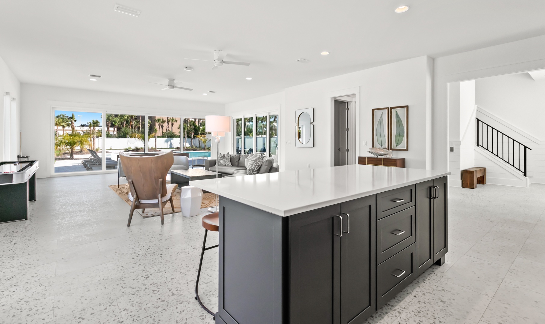 Picture yourself in this stunning open-concept living space, where the modern kitchen island invites you to gather while sunlight pours through floor-to-ceiling windows revealing your private pool and lush palm-fringed patio beyond.
