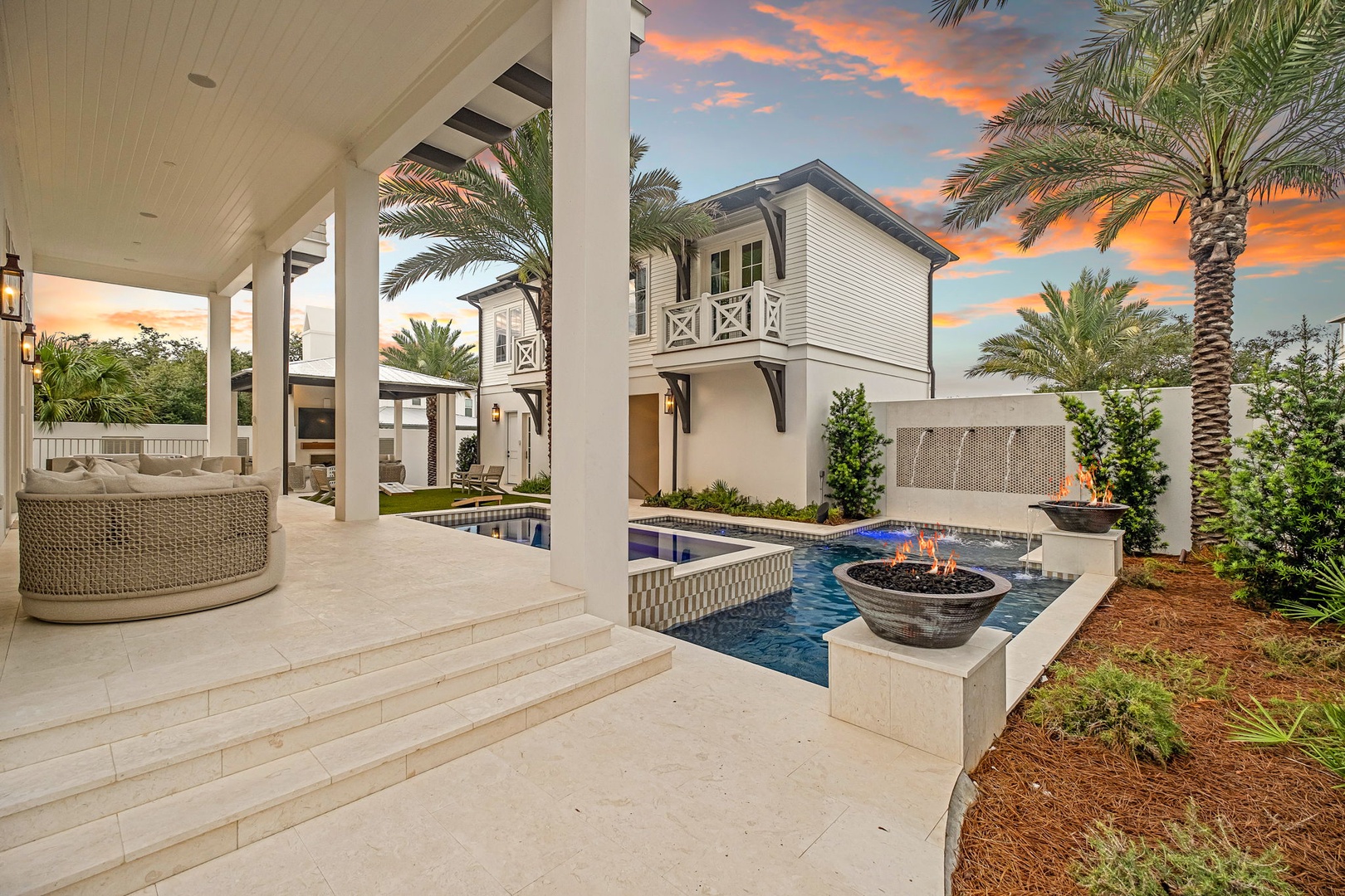 A luxurious backyard retreat showcasing a sparkling pool with a contemporary fire feature beneath a vibrant sunset sky, framed by swaying palms and elegant covered patio space perfect for relaxation.