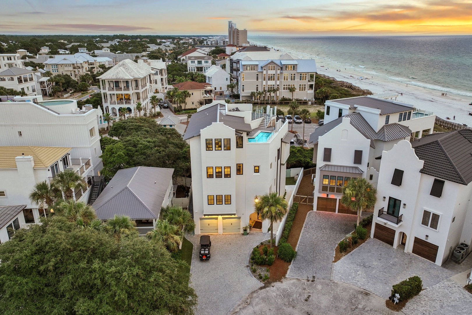 Aerial view captures a stunning coastal neighborhood with modern beach homes, lush landscaping, and pristine sandy shores stretching along the Gulf waters at sunset.