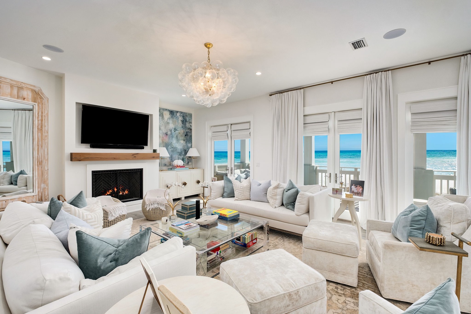 Sink into plush sofas and let the ocean view captivate you—your private coastal retreat features a cozy fireplace, elegant chandelier, and floor-to-ceiling windows framing endless turquoise waters.