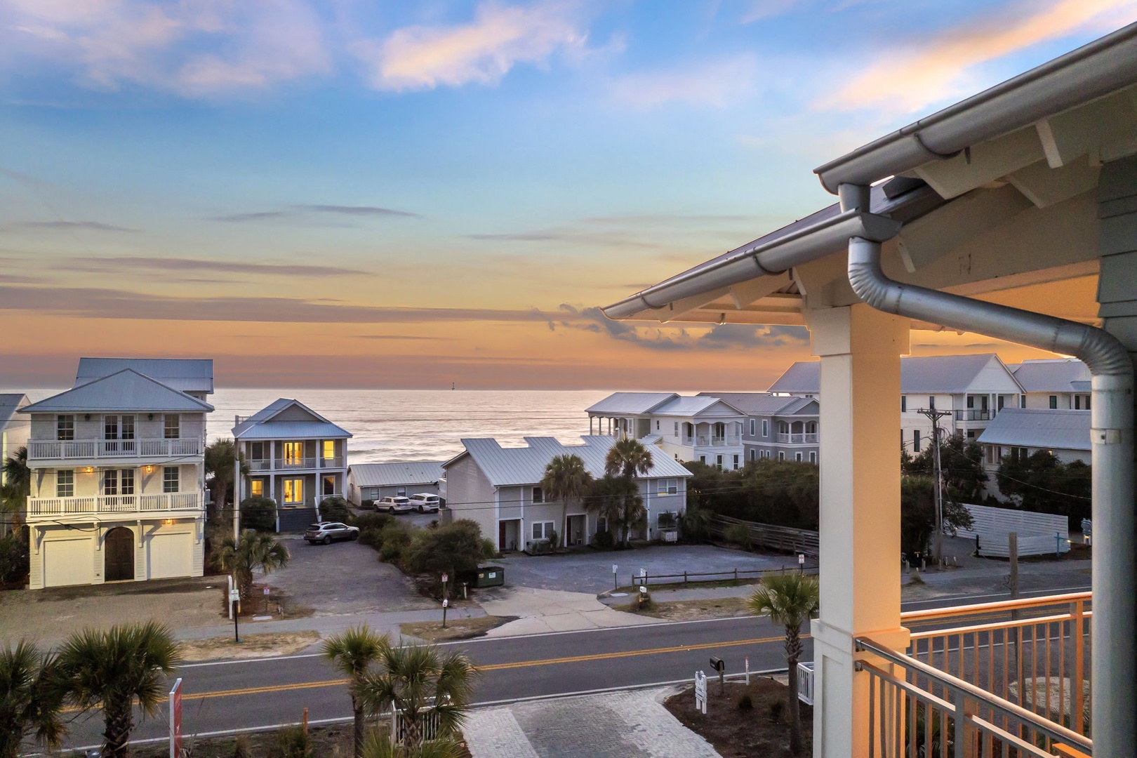 Coastal neighborhood view from elevated balcony, featuring beach houses and ocean glimpses at sunset.