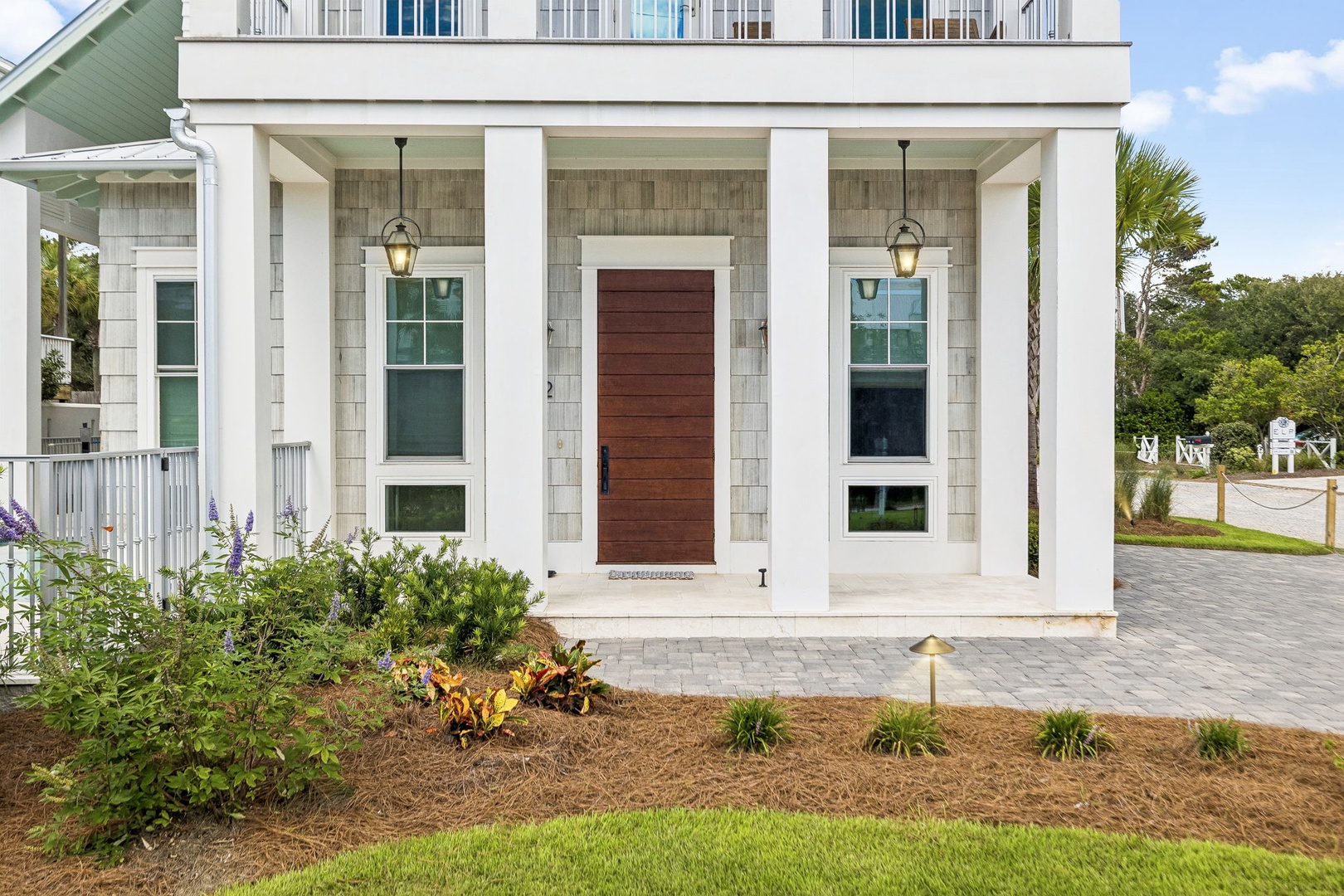 The property welcomes guests with classic coastal architecture, featuring clean white columns, natural wood accents, and professional landscaping surrounded by lush greenery and palm trees.