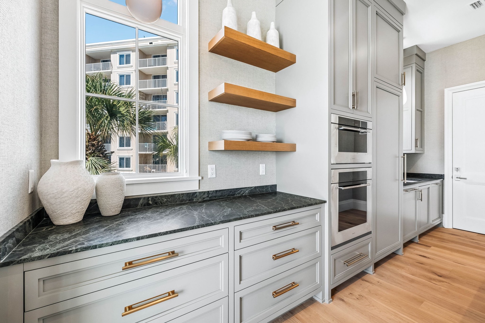 Your kitchen is bathed in natural light through tall windows with tropical palm views, and features sleek dark stone counters, warm wood shelving, and stainless steel double ovens ready for home cooking.