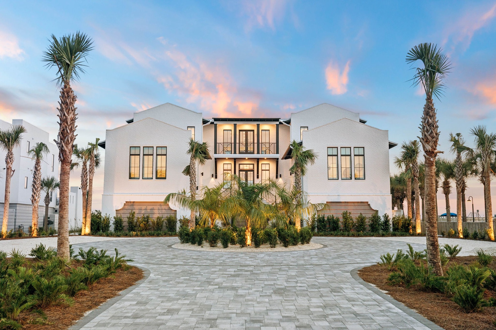A striking beachfront estate framed by swaying palms and tropical landscaping, welcoming guests to their coastal retreat at sunset.