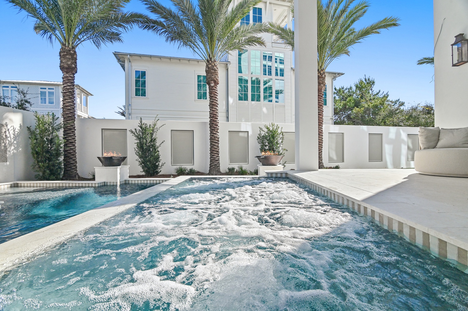 The property features an elegant outdoor oasis with a private hot tub and swimming pool, framed by towering palms and modern coastal architecture under a bright blue sky.