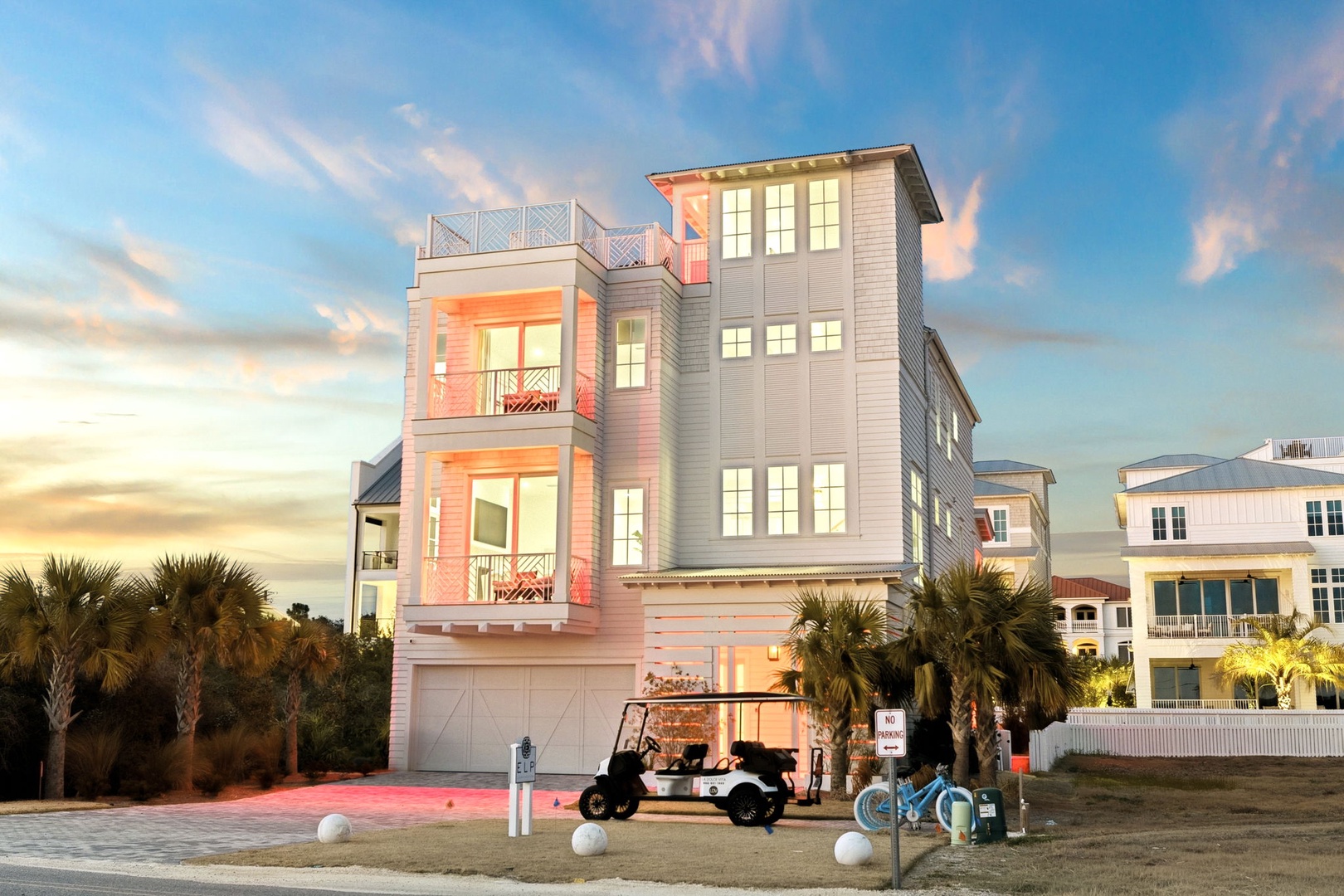 Modern three-story coastal home with rooftop deck and garage, featuring contemporary design with warm evening lighting in a beachfront neighborhood.