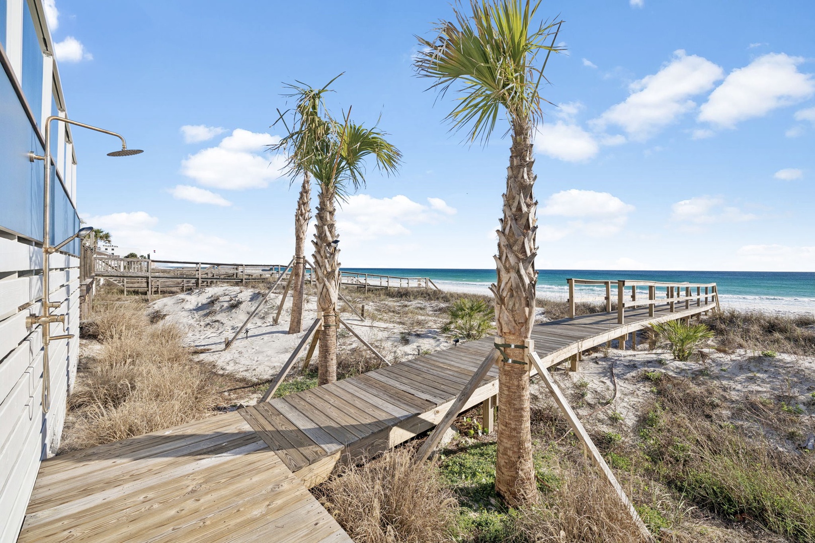 Wooden boardwalk winds through coastal dunes to pristine white sand beach with swaying palm trees and turquoise waters.