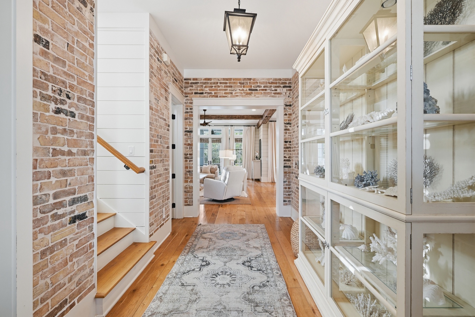 Step into a welcoming hallway where rustic exposed brick meets coastal elegance. Your eyes will be drawn to the warm hardwood floors, elegant glass display cabinets, and charming lantern lighting that creates an inviting first impression.