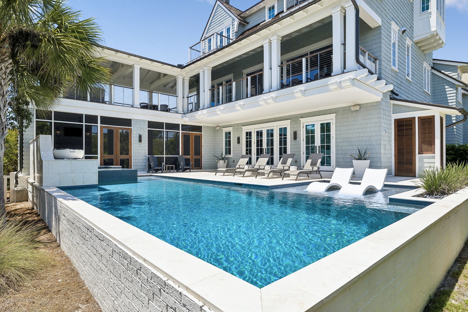 Elegant coastal property featuring an elevated pool deck with lounge seating, multiple balconies, and tropical landscaping under clear blue skies.