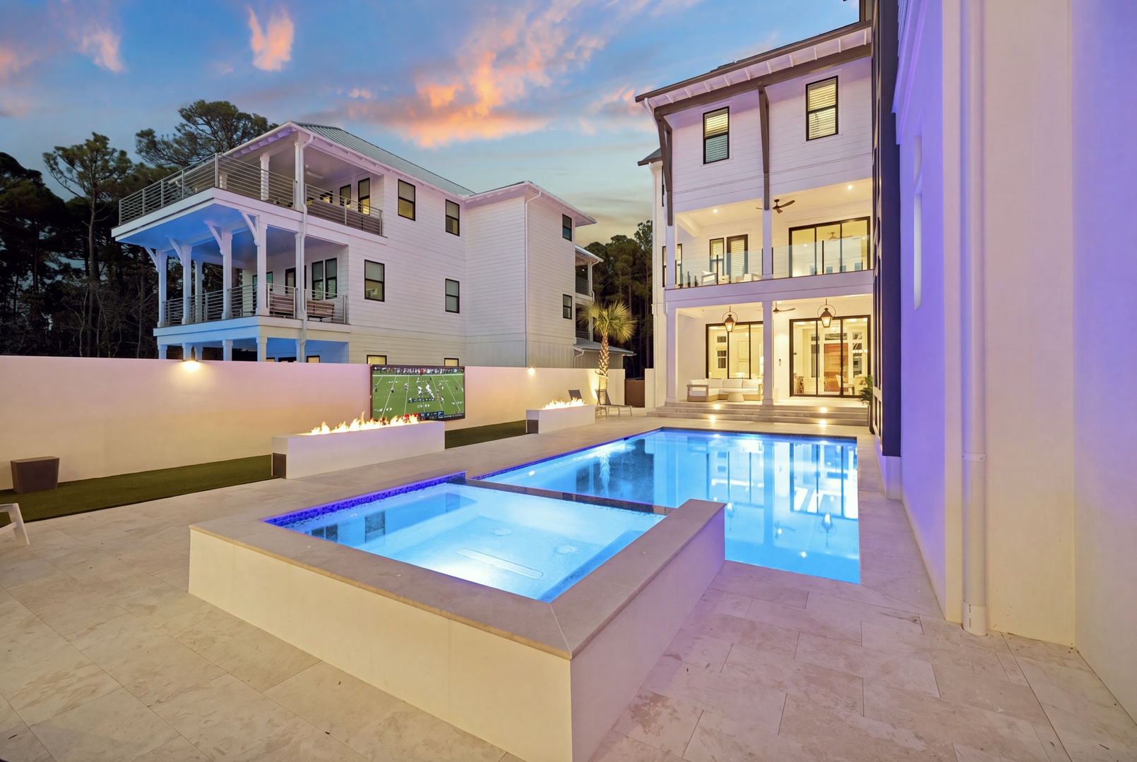 This stunning waterfront estate features a private pool, elevated spa, outdoor entertainment area with TV, and multiple balconies—all set against a backdrop of coastal beauty at twilight.