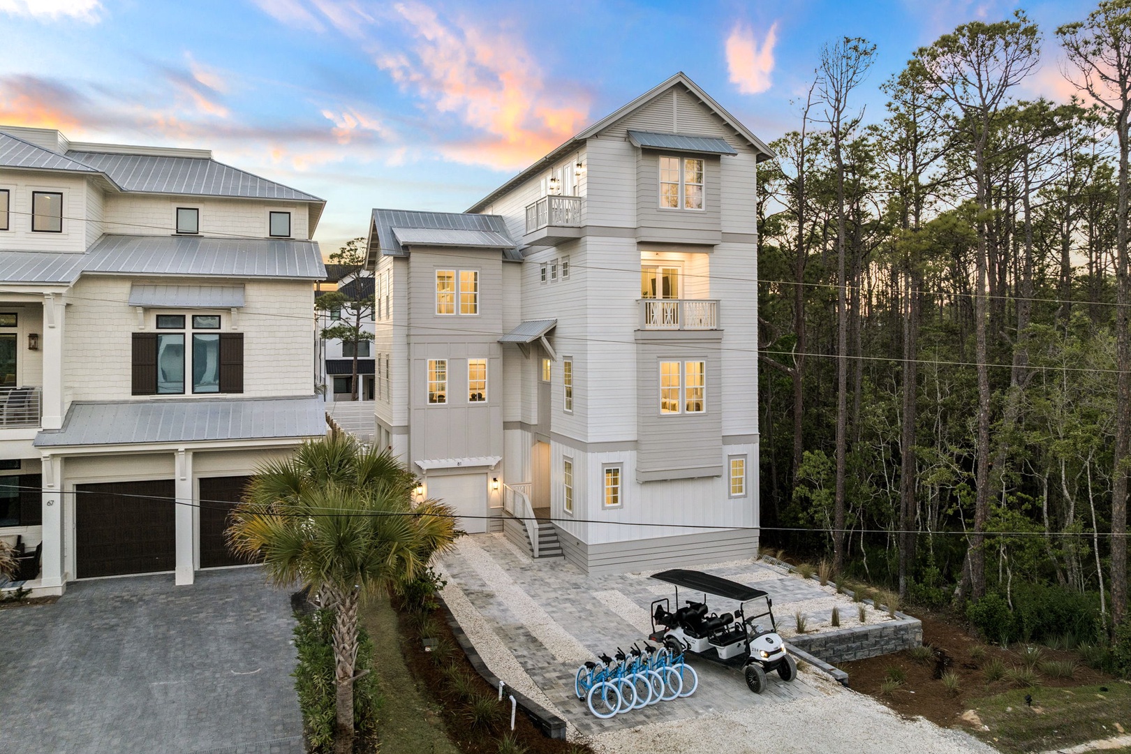Elegant multi-story beach house with illuminated windows set against a stunning sunset sky, surrounded by lush coastal vegetation and modern amenities.