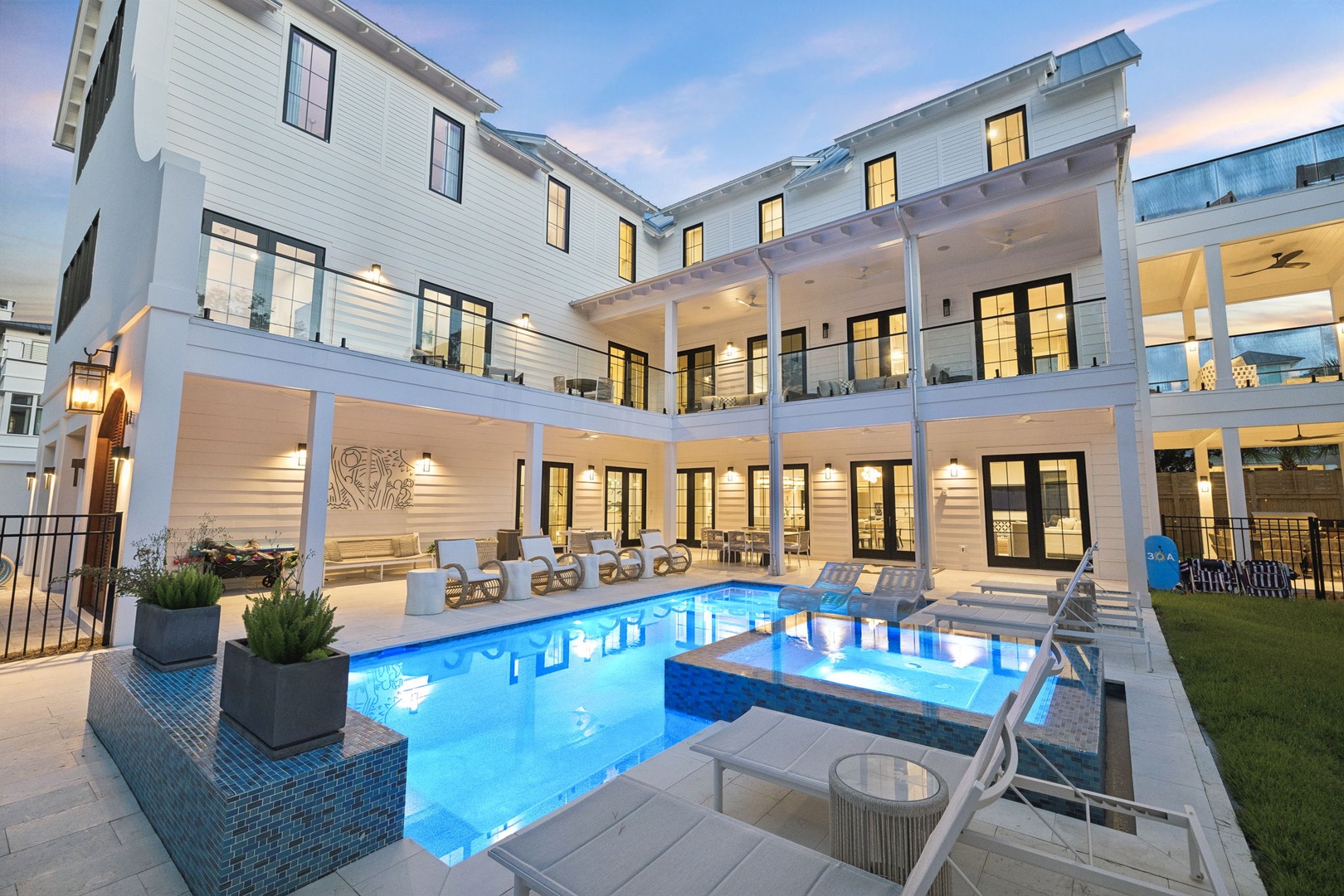 This modern white coastal property features a stunning pool with spa, multiple covered porches, and contemporary architecture. Captured as evening approaches, with warm interior lighting glowing through large windows.