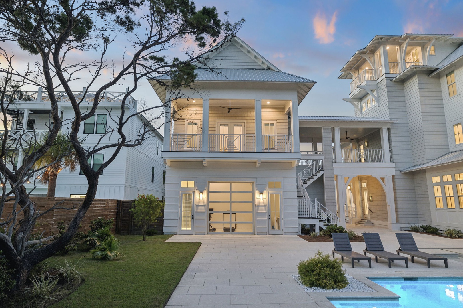 A stunning coastal retreat captured at twilight, where modern elegance meets beachside charm. The property glows warmly against the evening sky, with a sparkling pool and loungers ready to welcome guests to paradise.