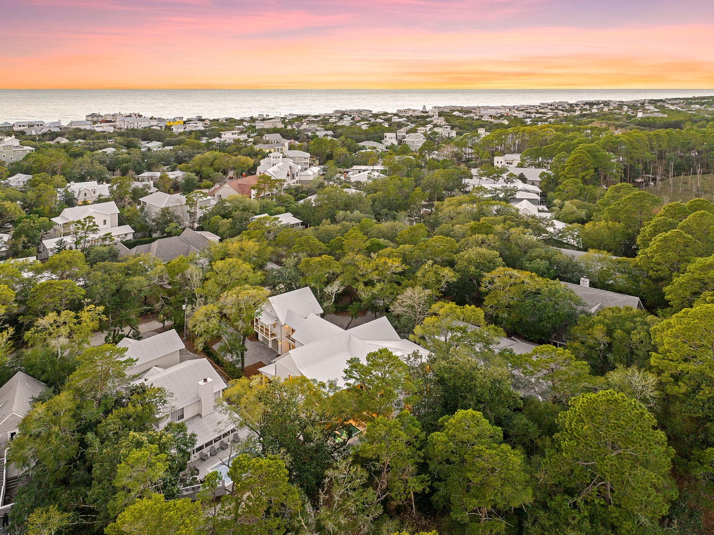 Aerial view showcases this coastal neighborhood nestled among lush trees, with the beach and ocean nearby during golden hour.