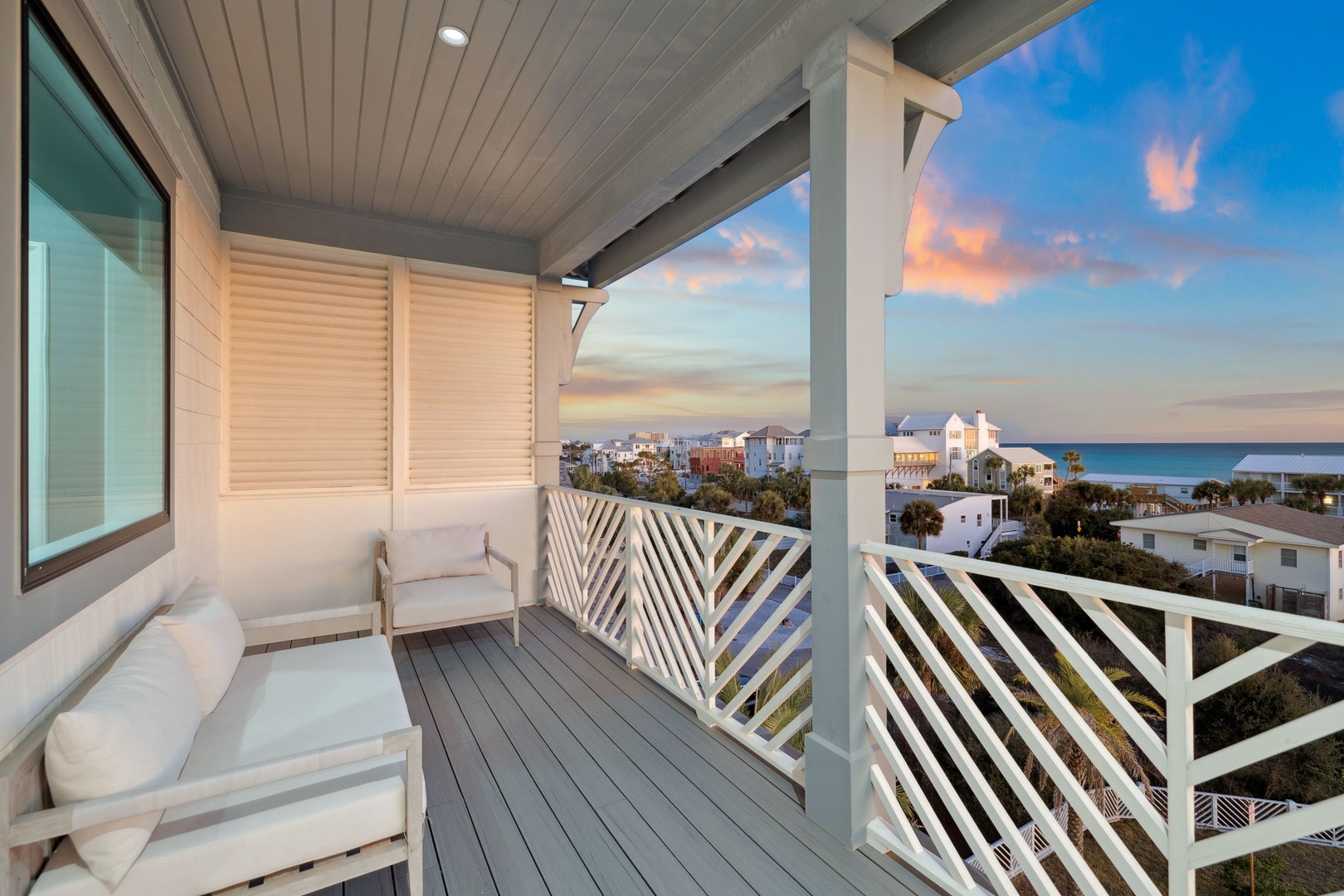 Unwind on your private balcony with comfortable seating as vibrant sunset colors paint the sky over coastal homes and ocean beyond.