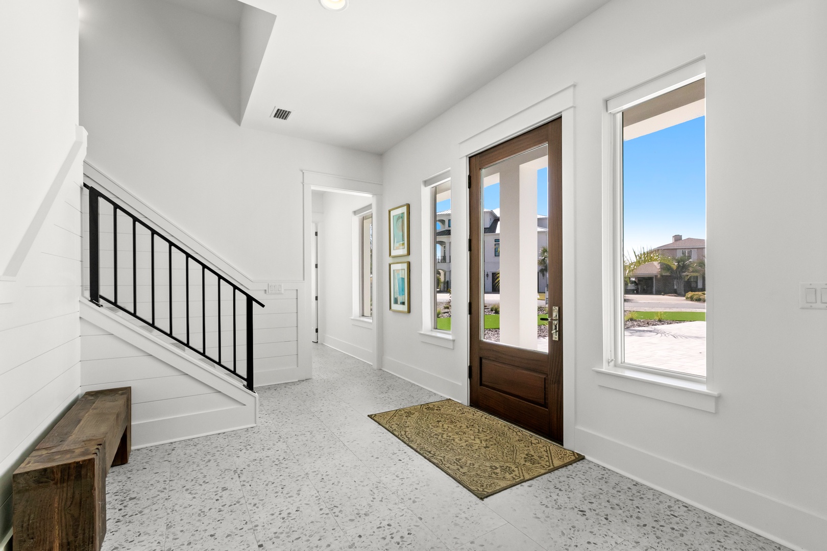 A bright, airy entryway greets you with clean white walls, modern staircase, and natural light streaming through the glass door and windows. The terrazzo-style flooring adds a contemporary touch to this welcoming space.