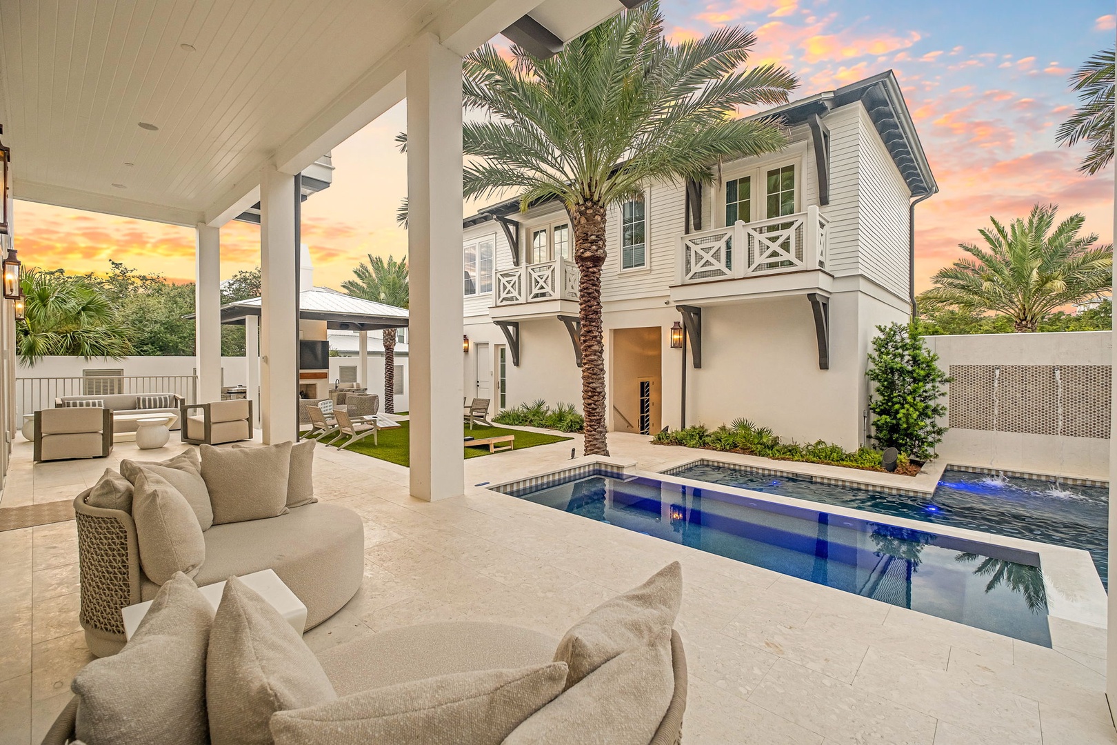 Your private oasis awaits beneath a covered patio where plush seating frames a sparkling pool, swaying palms, and stunning sunset skies painted in gold and coral.