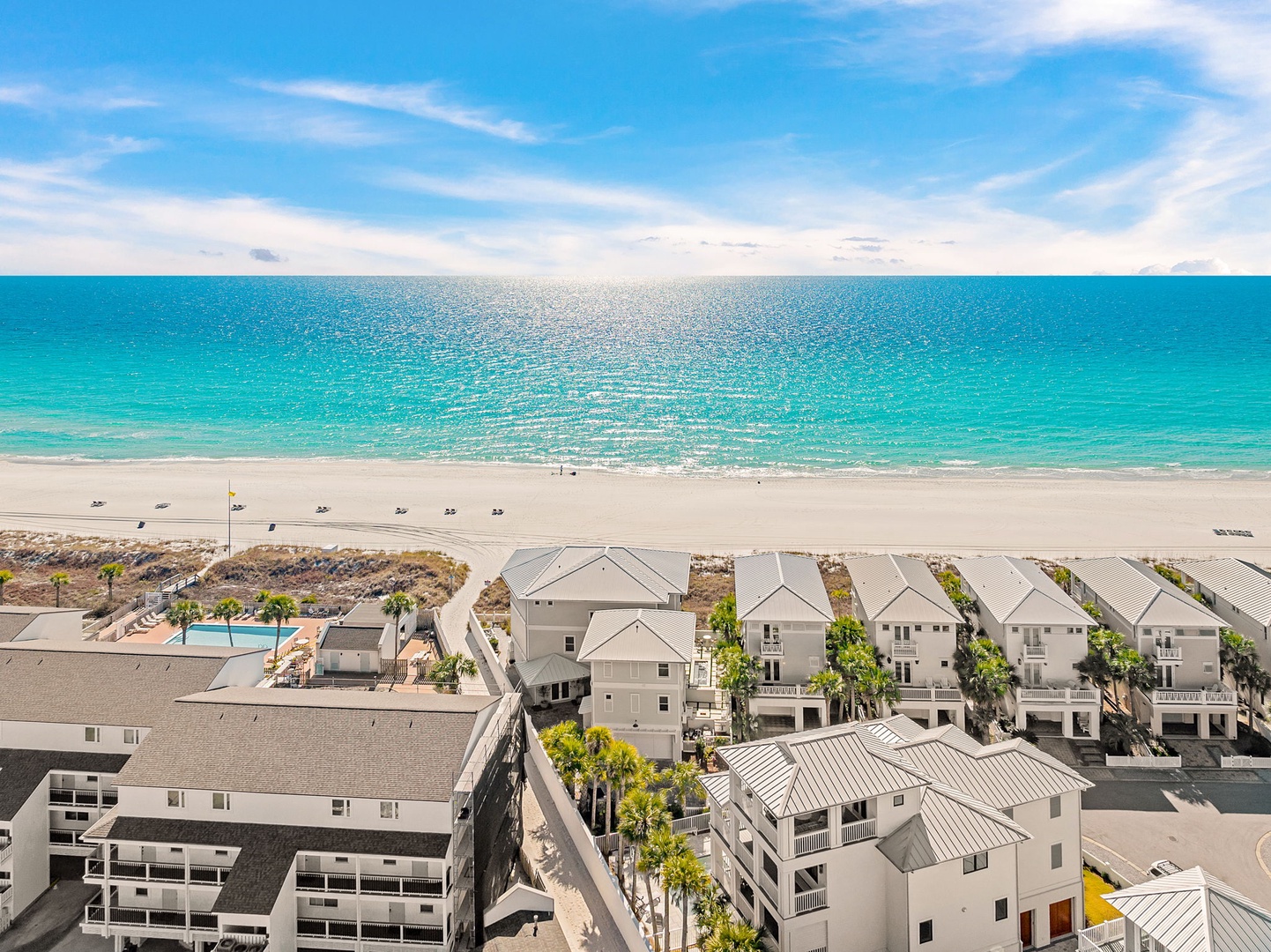 Elevated aerial view showcasing a pristine beachfront resort community with direct access to turquoise waters and white sand, featuring modern coastal architecture and a sparkling pool area steps from the shore.