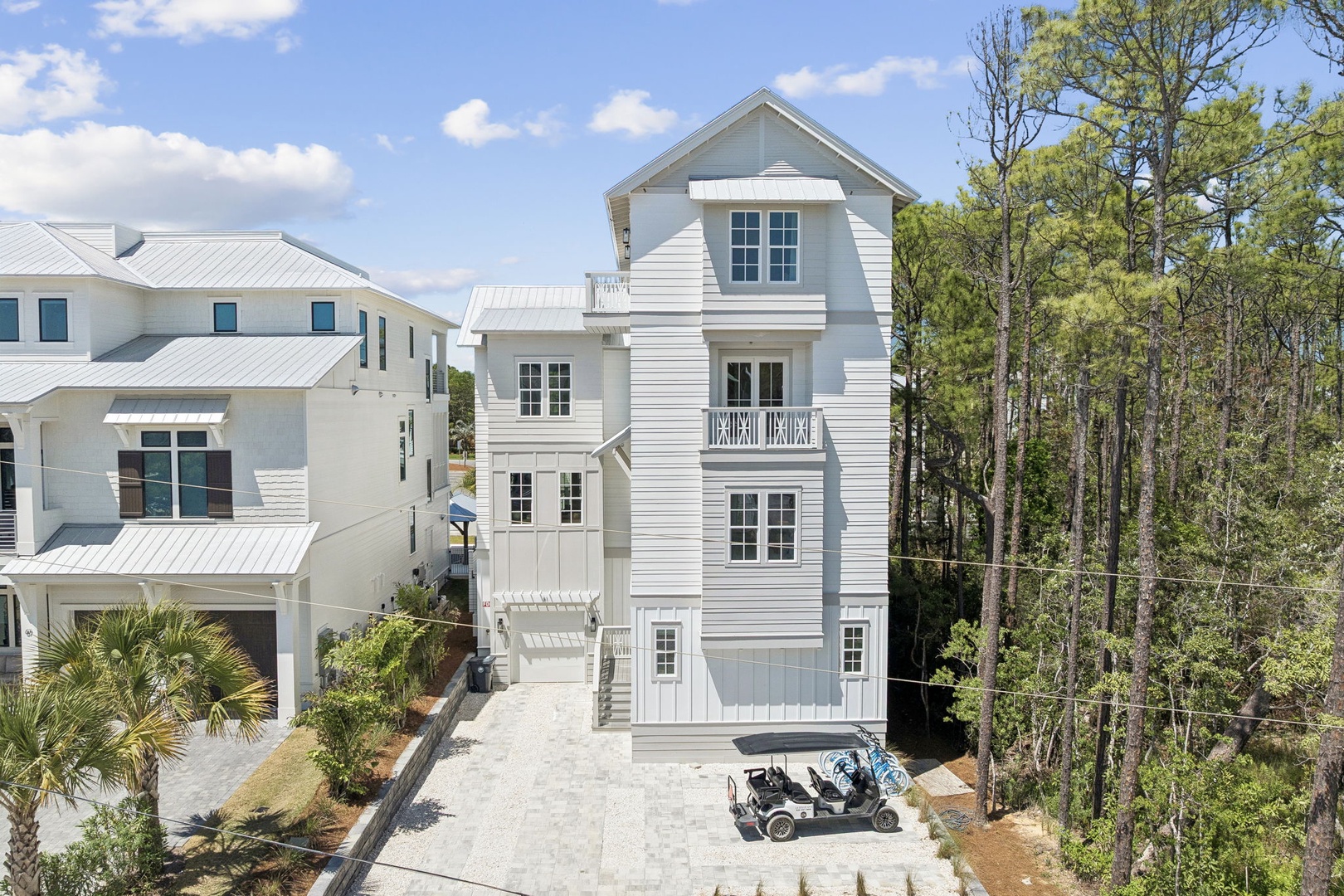 Modern coastal property featuring a multi-story white home with metal roof, nestled among lush trees in a quiet neighborhood setting.