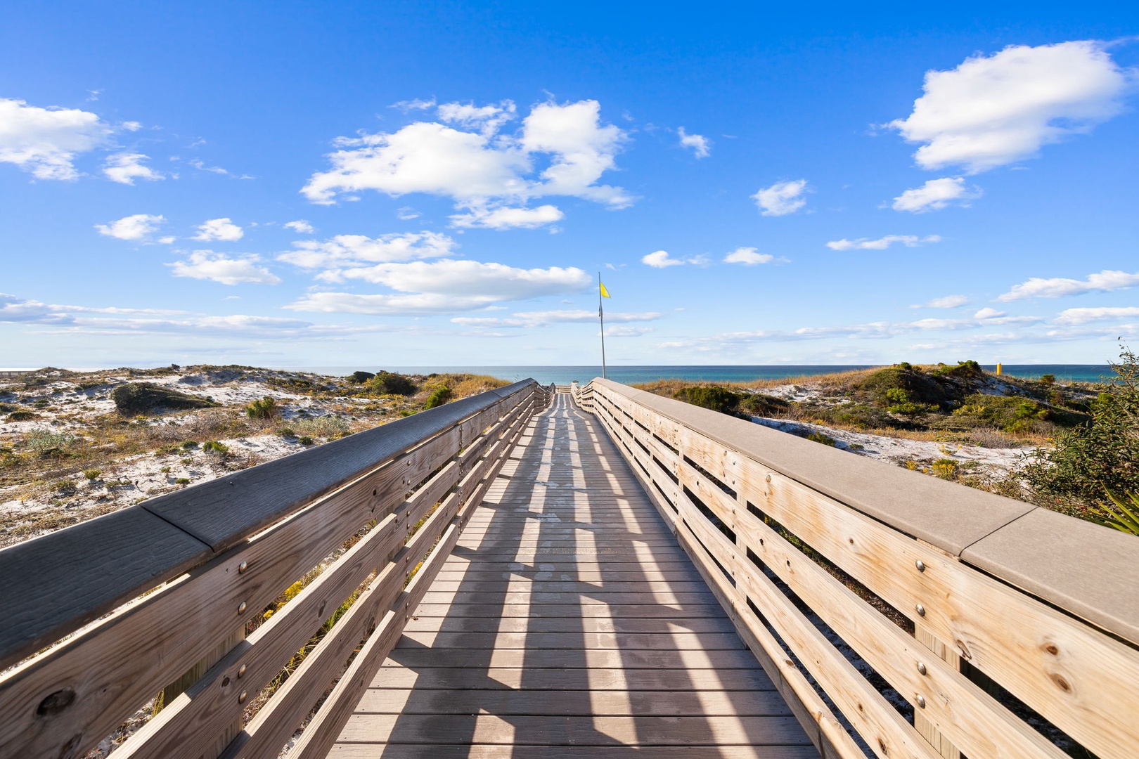 Wooden boardwalk leads through coastal dunes toward pristine beach under brilliant blue skies with scattered white clouds.