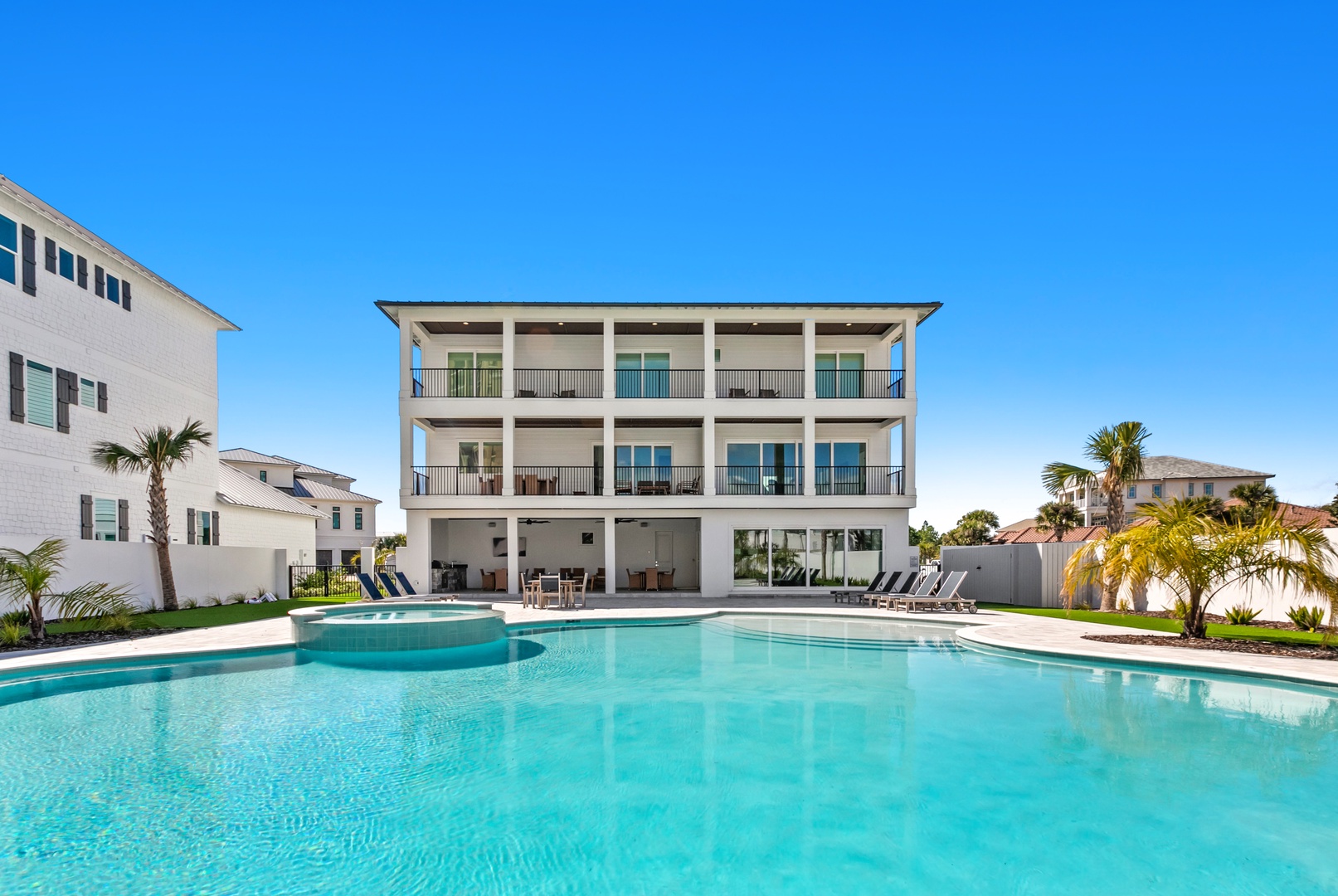 The property features a spacious modern exterior with multiple balconies overlooking a stunning resort-style pool, perfect for soaking up sunshine in a tropical setting surrounded by palms.