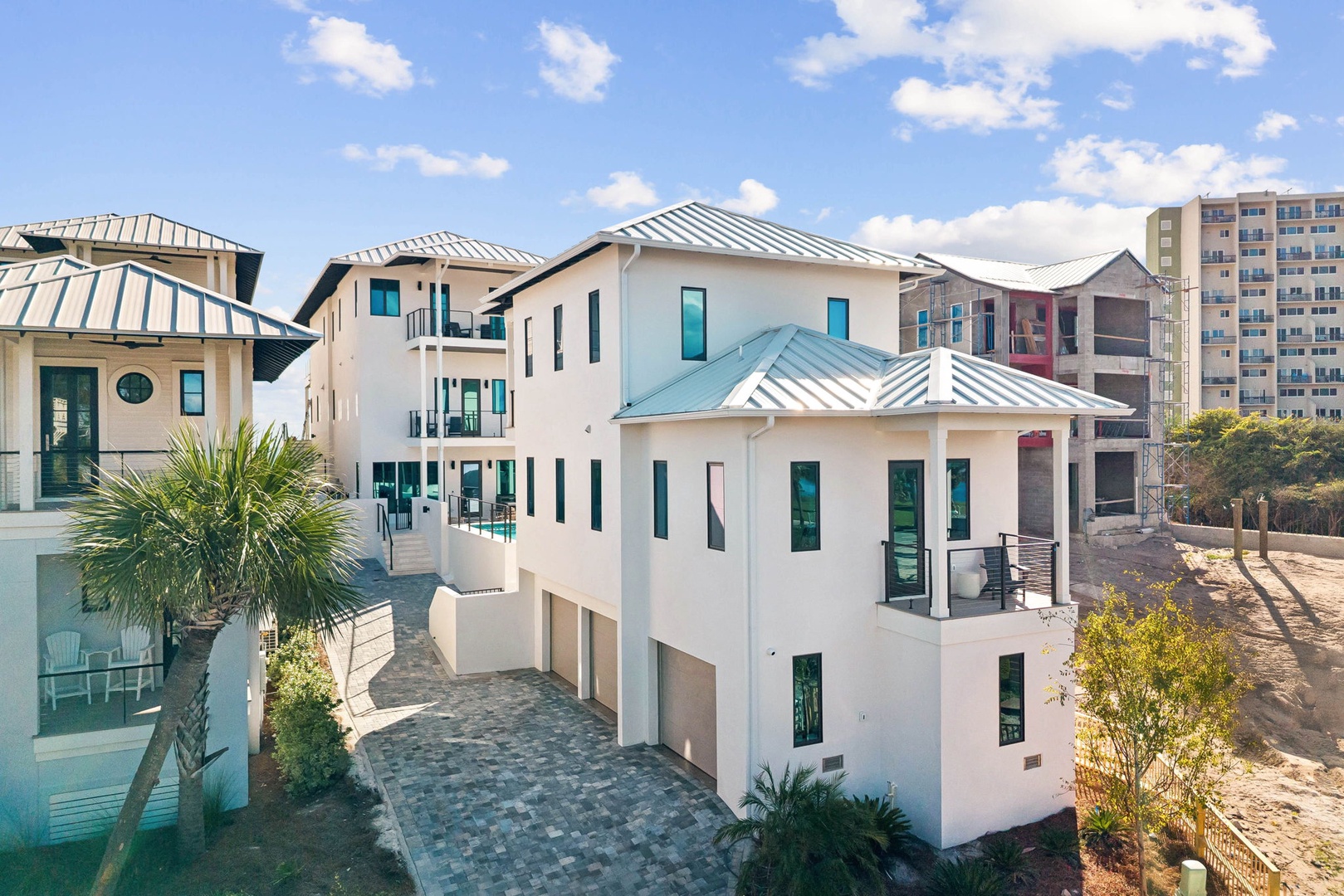 Modern coastal vacation home featuring bright white stucco exterior, metal roof, and multiple covered balconies with coastal landscaping, nestled in a beachfront neighborhood.