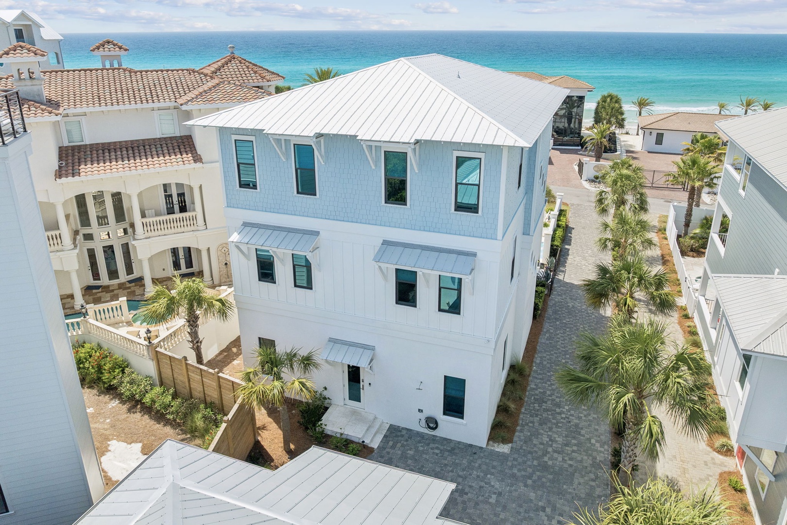 Modern three-story coastal home with white metal roof and turquoise accents, surrounded by tropical palms just steps from pristine turquoise waters.