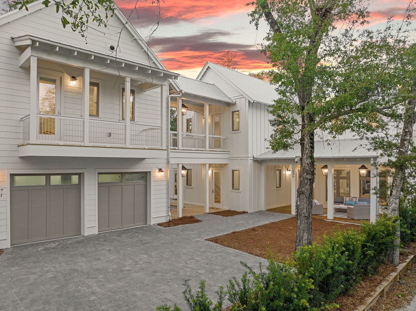Modern coastal-style home with garage parking and covered porches, set against dramatic sunset skies and mature oak trees.