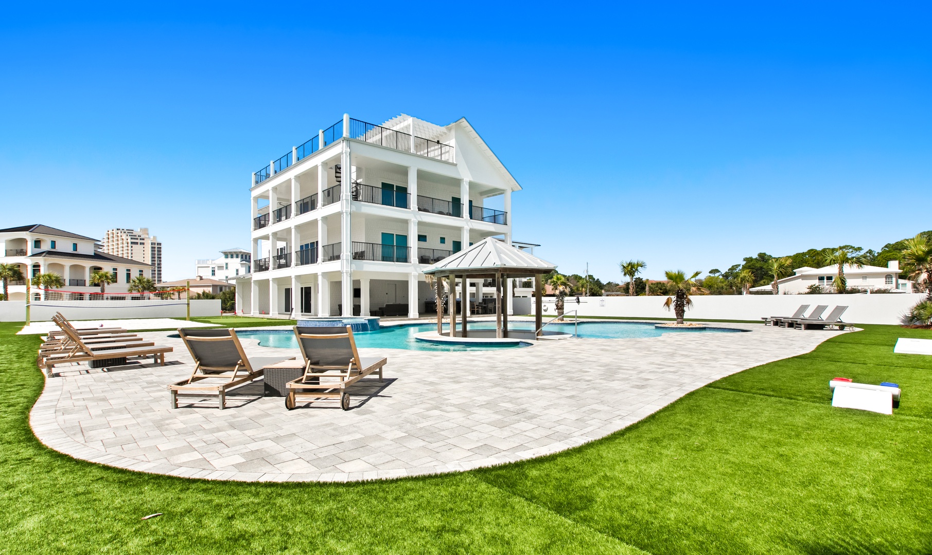 A stunning multi-level vacation home features resort-style grounds with a sparkling swimming pool, covered pavilion, and multiple sun loungers set on a curved paver patio, all framed by lush green lawn and swaying palm trees.