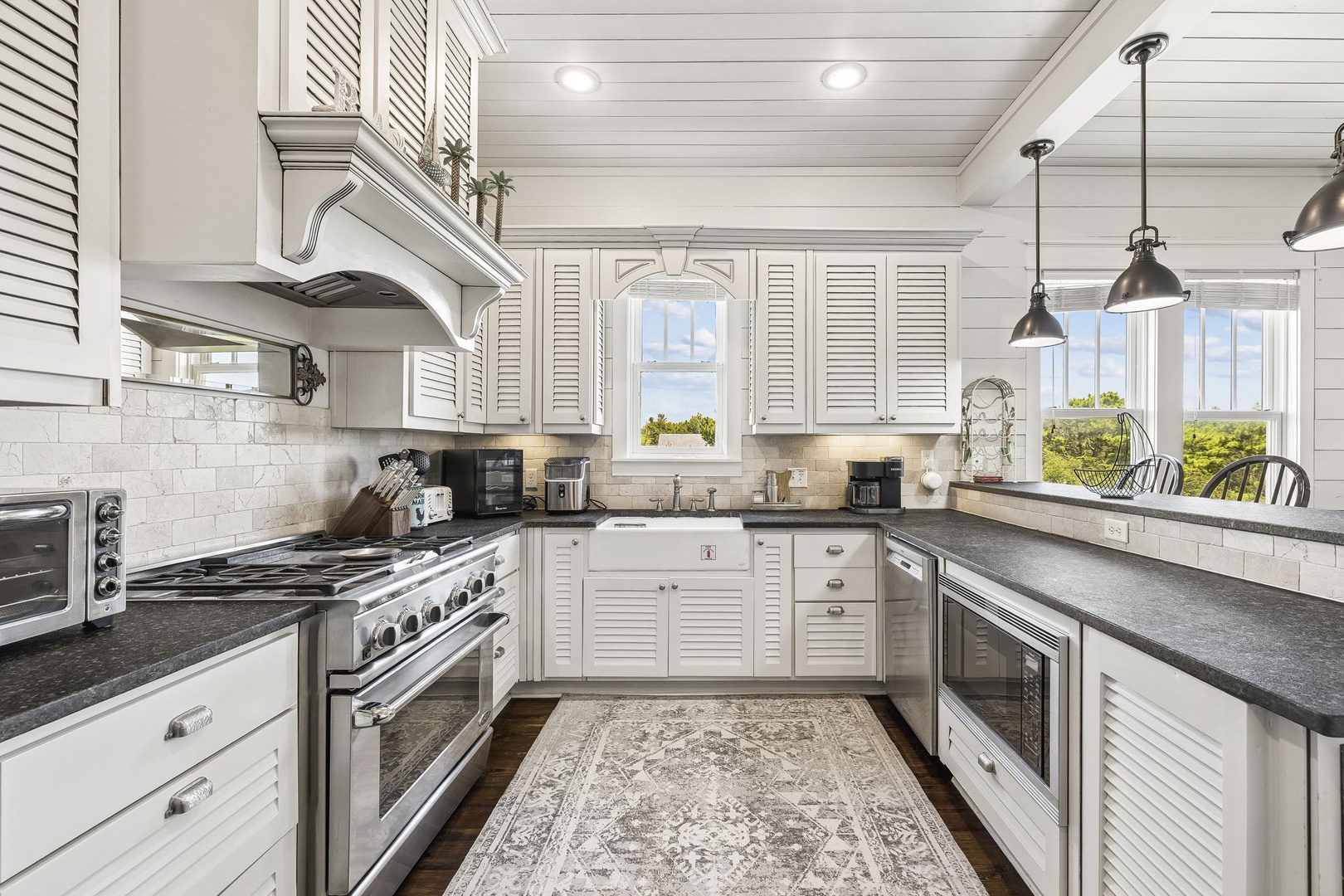 A beautifully designed kitchen with bright white shiplap walls, louvered cabinetry, and sleek dark countertops awaits your culinary adventures. Large windows frame lush greenery, filling the space with natural light.