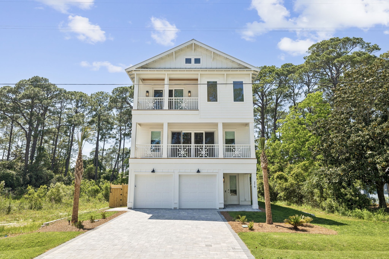 A three-story coastal home with dual balconies and two-car garage, surrounded by mature trees and landscaped grounds in a quiet residential area.