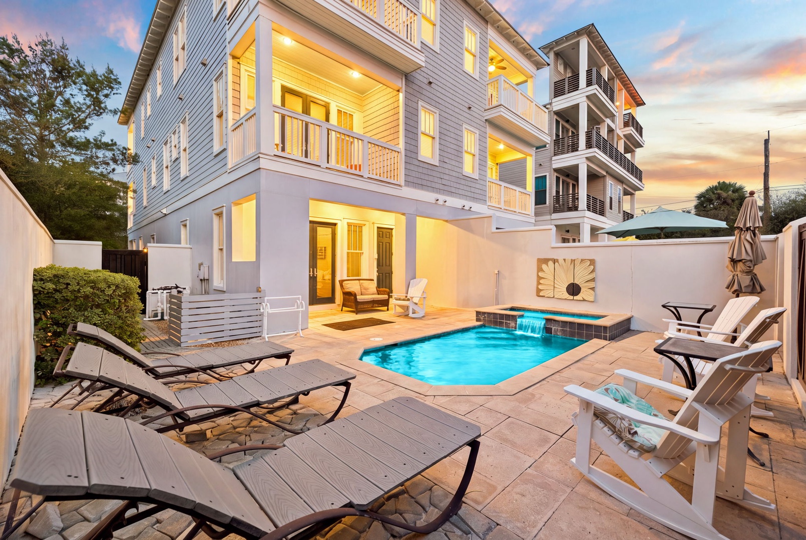 A stunning twilight view of this modern coastal property reveals your private outdoor sanctuary, complete with a sparkling pool, inviting loungers, and multiple seating areas perfect for unwinding under the evening sky.