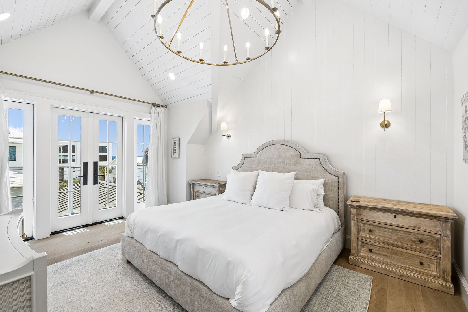 Your serene coastal bedroom features vaulted shiplap ceilings, elegant brass lighting, and a plush upholstered bed dressed in crisp white linens. French doors open to your private balcony, inviting soft breezes and natural light.