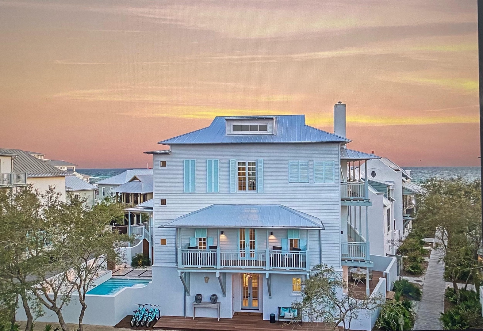 Stunning coastal retreat with blue shutters and private pool, framed by a golden sunset sky over the ocean horizon.