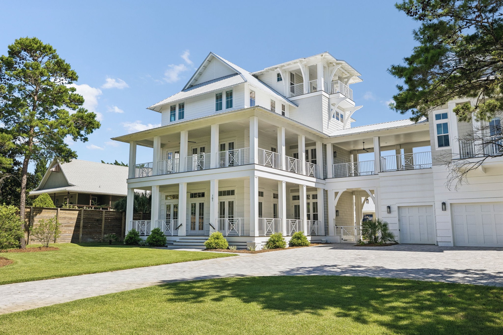 This impressive coastal-style property features a grand white facade with spacious wraparound porches and multiple private balconies, surrounded by landscaped lawns and mature pines.