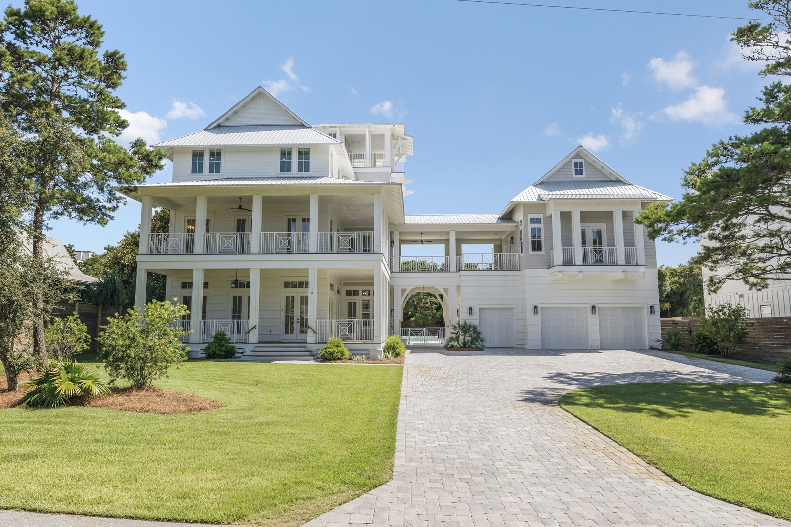 A stunning coastal property welcomes guests with elegant Southern architecture, featuring wraparound porches and spacious balconies surrounded by lush greenery and mature pines.