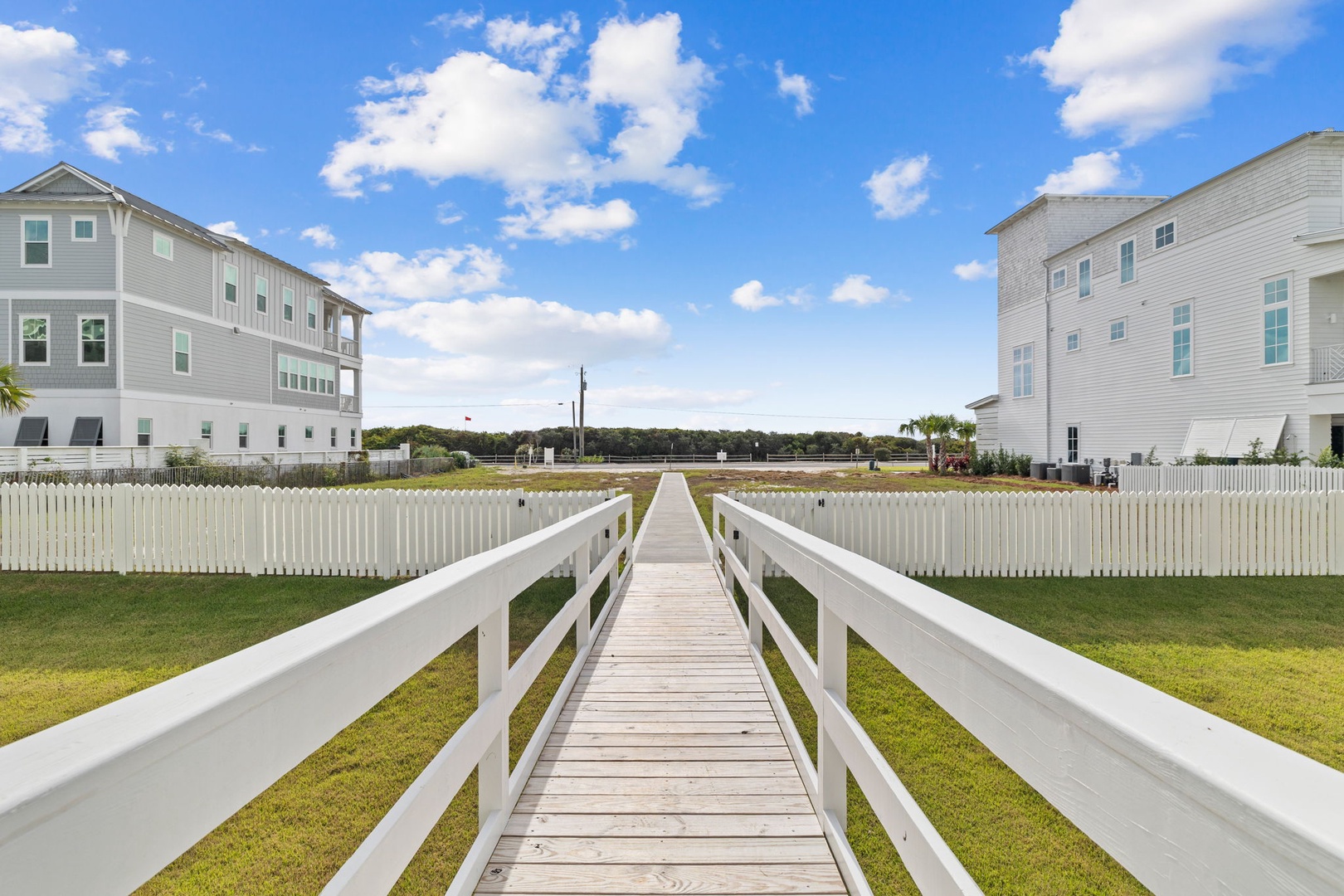 A charming wooden boardwalk leads through a coastal neighborhood of elegant beach houses under brilliant blue skies.