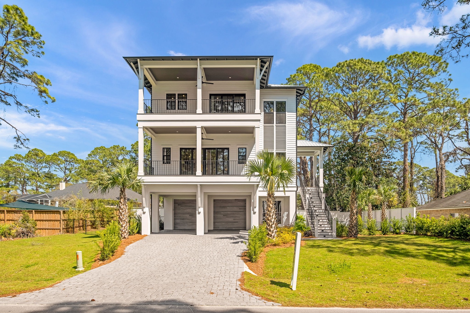 Modern three-story coastal home featuring covered balconies, palm-lined driveway, and spacious lawn surrounded by mature trees.