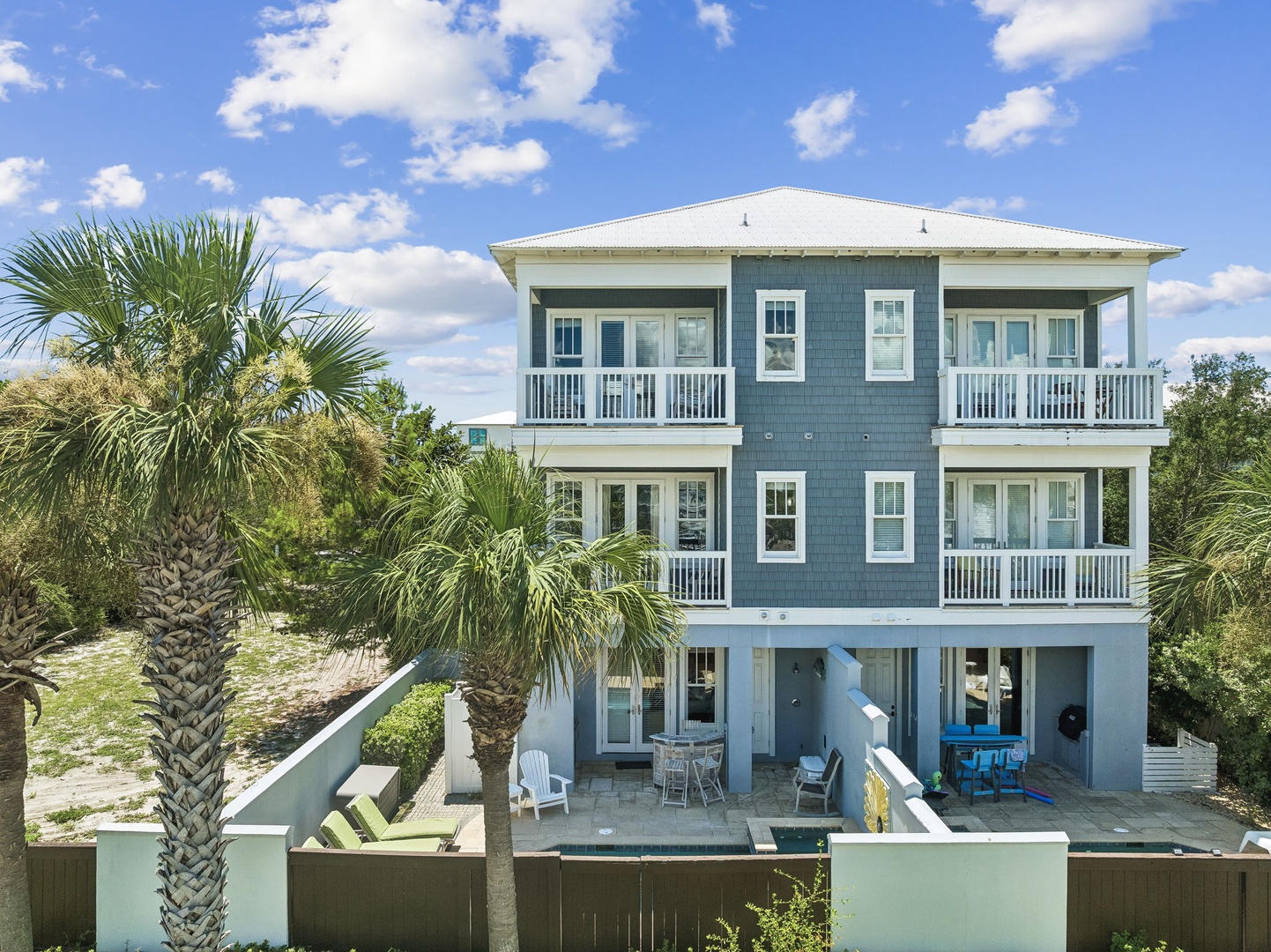 Coastal vacation home set in a tropical landscape with palm trees, featuring multiple balconies and ground-level outdoor living spaces beneath bright blue skies.