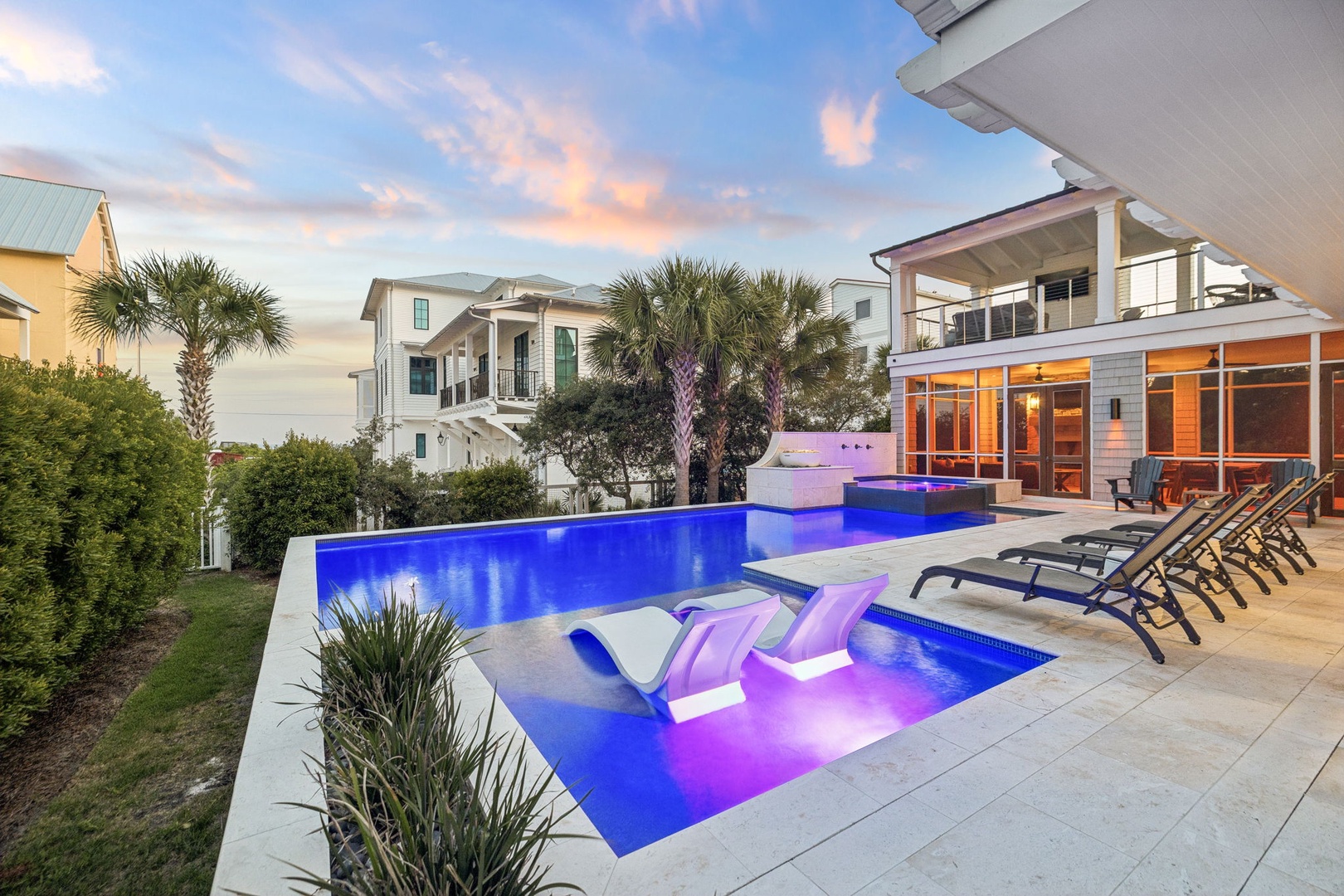 Luxurious coastal retreat featuring an illuminated infinity pool, integrated spa seating, spacious sun deck, and modern architecture perfectly framed by palm trees and neighboring beach homes.