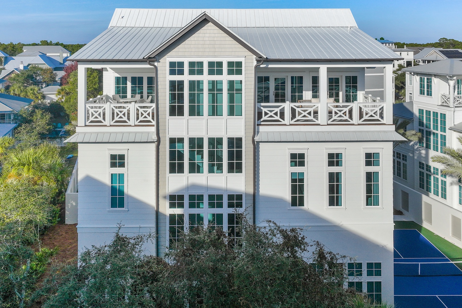 Modern coastal vacation home featuring spacious white balconies with decorative railings, a metal roof, and floor-to-ceiling windows. The property is nestled in a tree-lined neighborhood near recreation courts.
