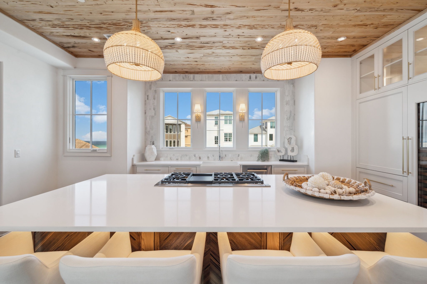 Gather around this bright kitchen island with coastal views through multiple windows. Natural wood ceilings and woven pendant lights create a warm, beachy atmosphere perfect for preparing meals together.