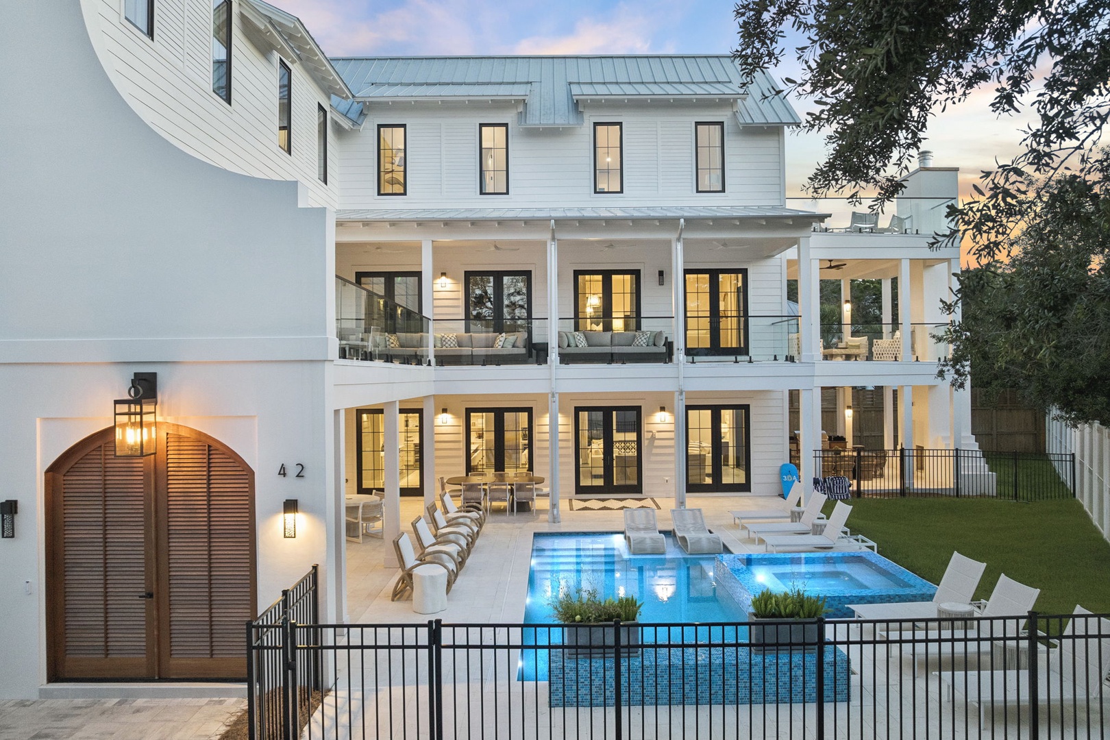 A stunning white coastal-style property featuring a sparkling pool, multiple covered balconies, and warm evening lighting that creates an inviting ambiance for your stay.