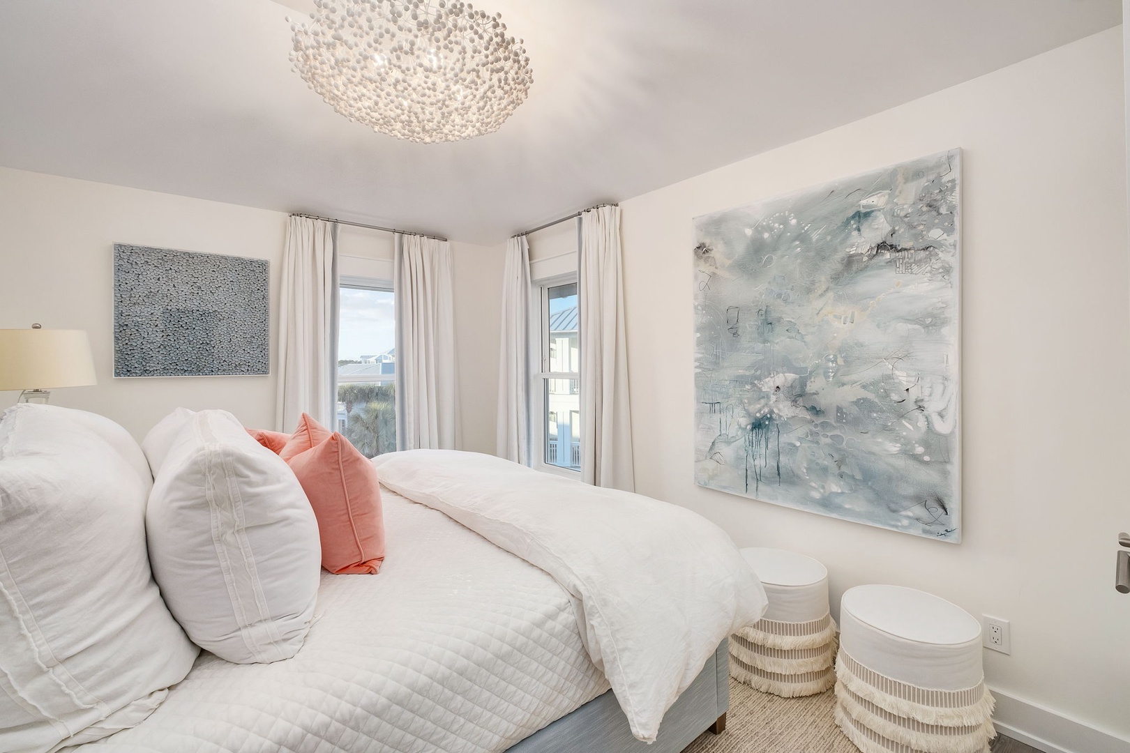 Sink into plush white linens beneath a sparkling crystal chandelier. Soft natural light and serene coastal artwork create your peaceful retreat with warm pops of coral accent.