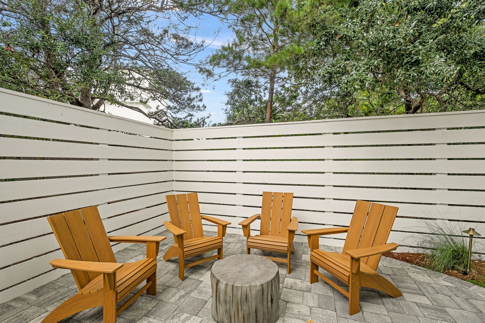 Your own secluded courtyard retreat, complete with Adirondack chairs and a natural wood fire table, surrounded by lush trees and modern privacy fencing.