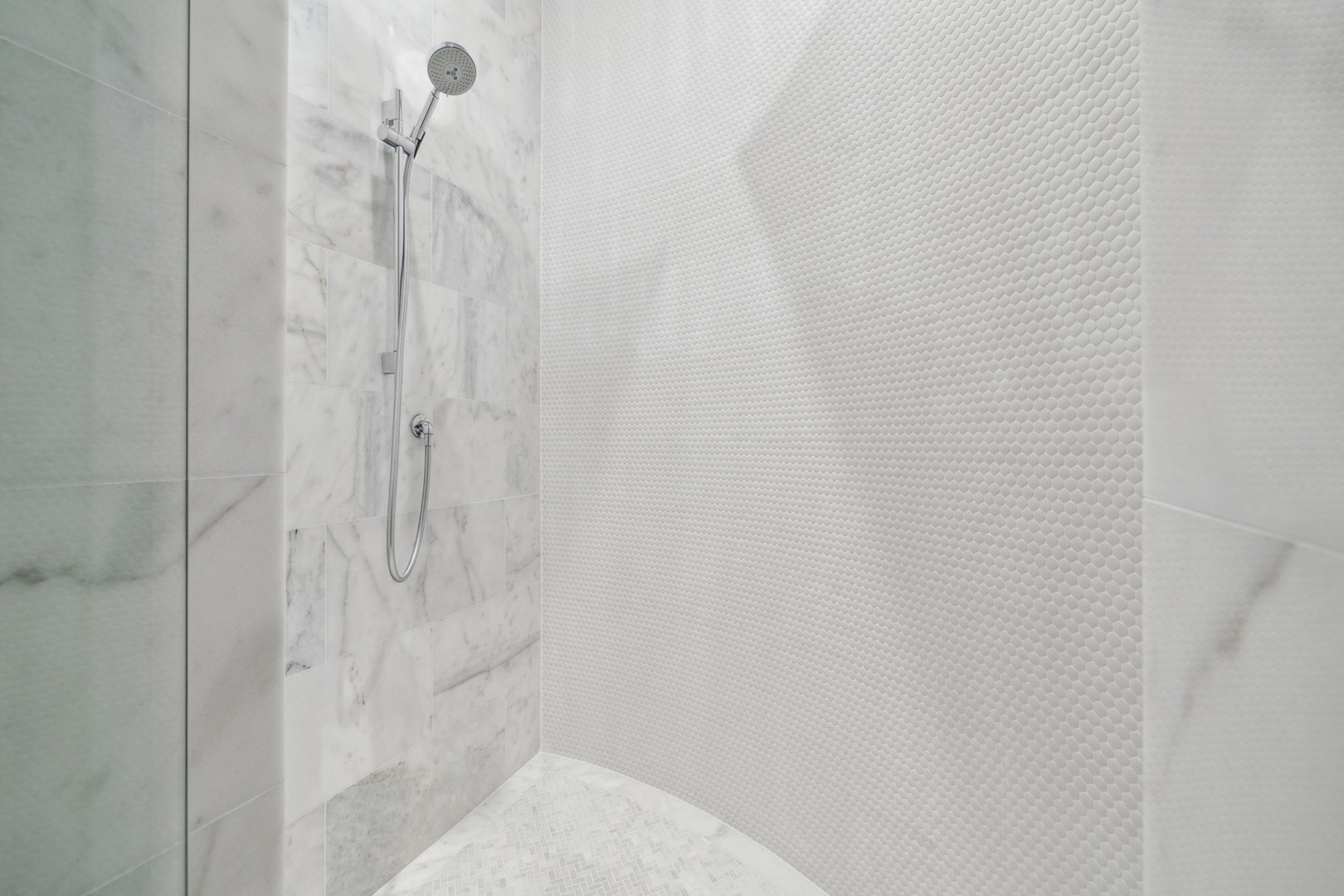 Step into your sleek, modern shower featuring elegant marble accents and pristine white tile. The adjustable handheld showerhead adds a touch of convenience to your daily refresh.