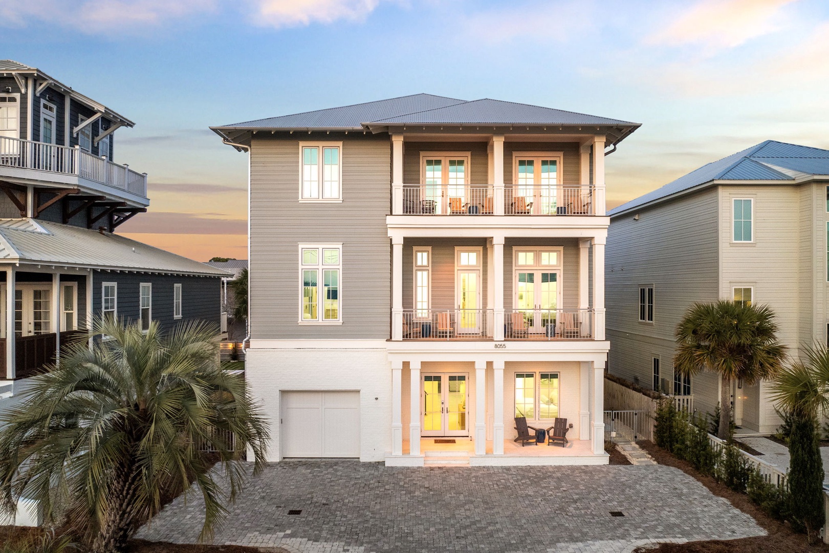 Modern three-story coastal home with elegant balconies and warm lighting, surrounded by palm trees in a beach community setting.