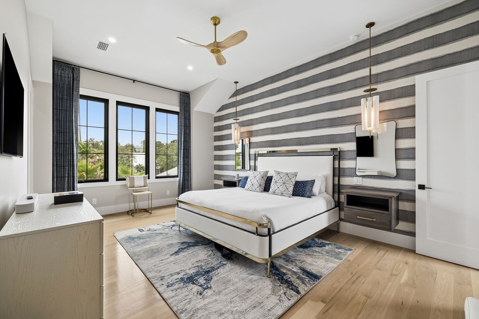 Your elegant master suite awaits with designer touches, natural light streaming through large windows, and a plush bed framed by striking striped accent wall and modern fixtures.