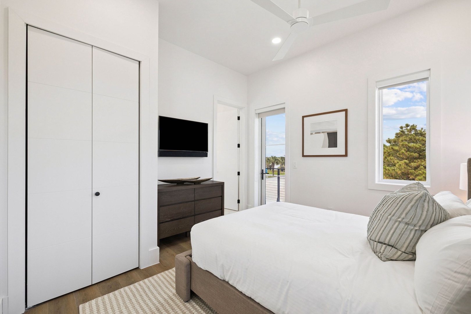 Your bright bedroom features crisp linens and balcony doors that open to coastal breezes, with sunny skies and natural scenery framing your peaceful retreat.