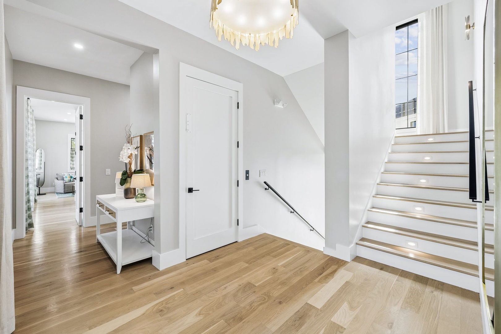 Step into your elegant entryway, where polished wood floors meet soaring ceilings and a sparkling chandelier overhead. Natural light pours in from the windows, guiding you up the modern staircase or through to the welcoming rooms beyond.